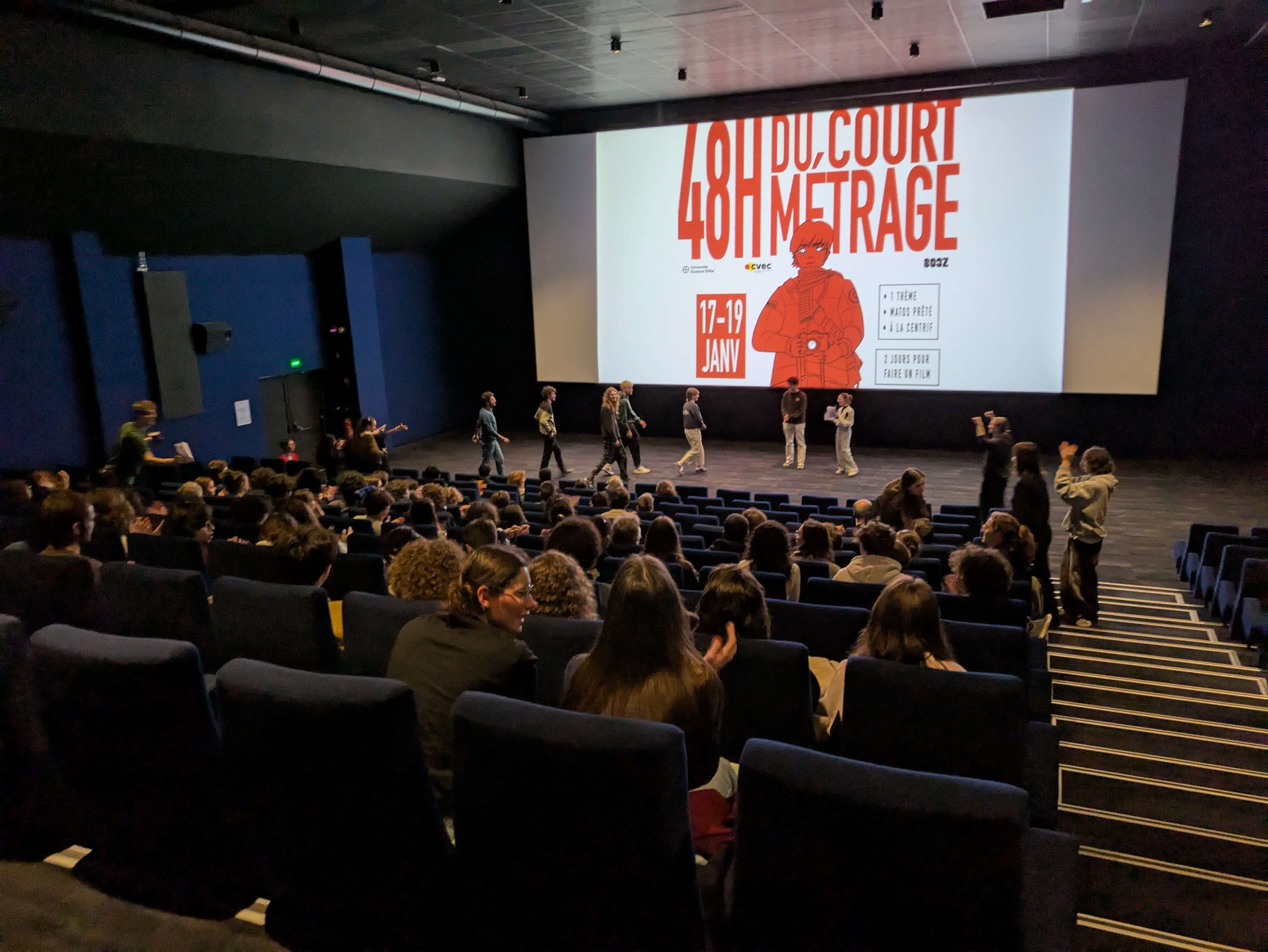 Le visuel de l'événement sur un écran de cinéma, avec des personnes sur scène face au public