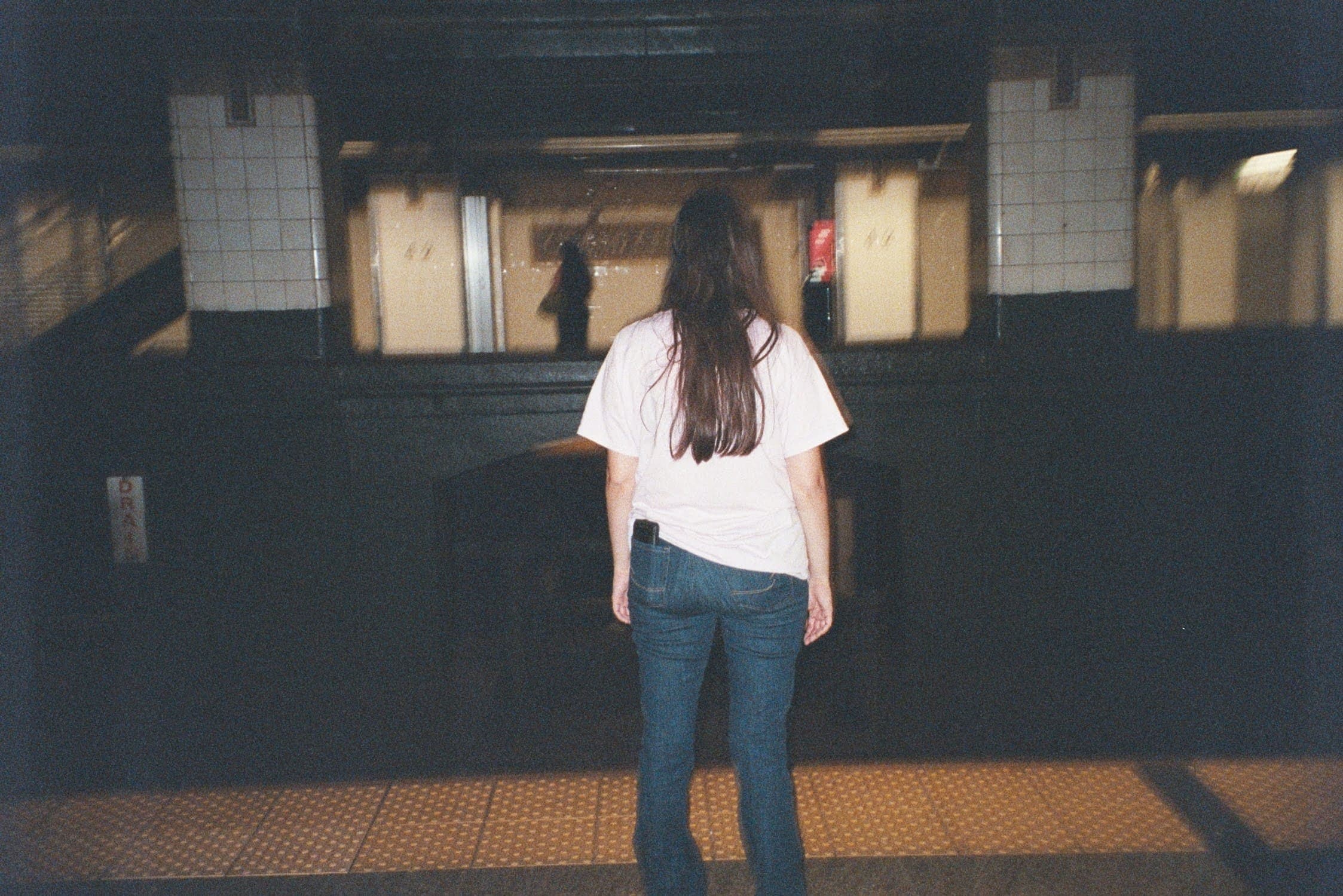 Une personne aux cheveux longs, vue de dos, debout sur un quai sombre du métro de New York